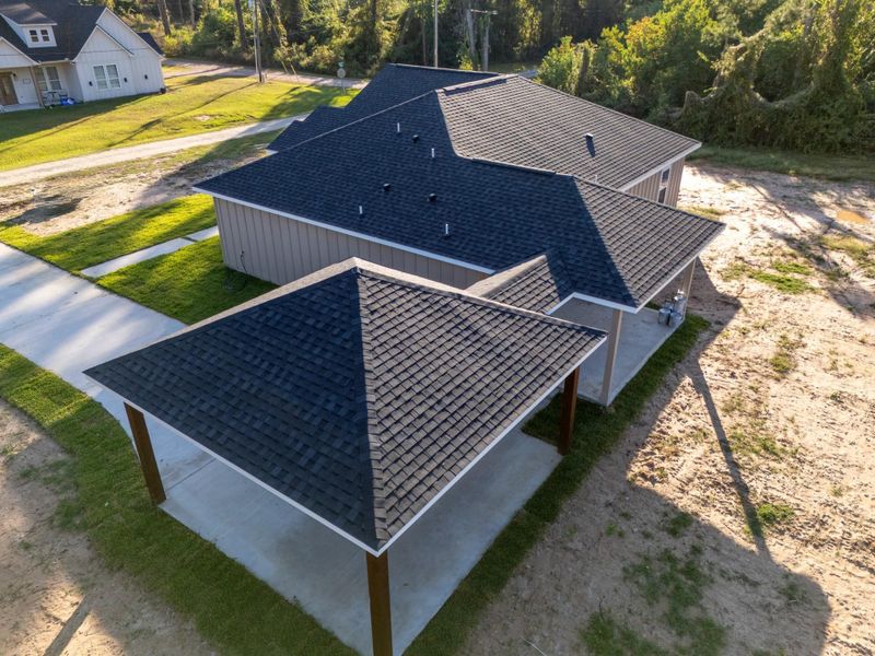 This photo shows a modern, single-story home with a dark shingle roof and a covered carport. The property is surrounded by a mix of grass and dirt patches, set in a quiet, wooded area.