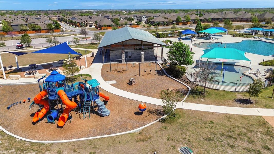 This photo showcases a vibrant community park featuring a colorful playground with slides, a covered picnic area, and a large swimming pool with shaded areas. It's an ideal spot for family activities and leisure, surrounded by a residential neighborhood.