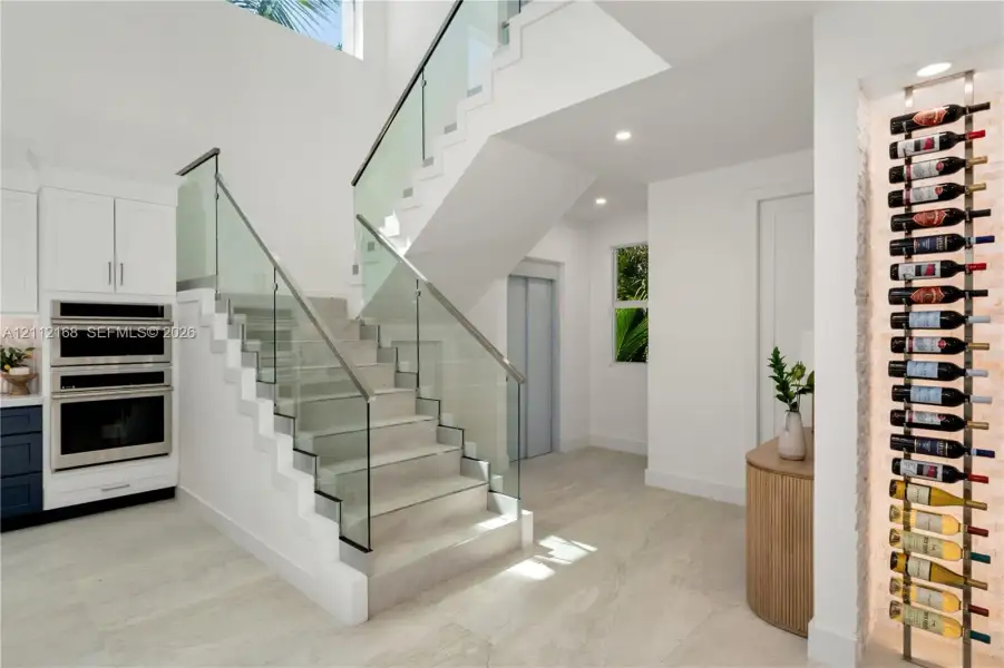 Open-riser staircase featuring a glass-panel railing, built-in stainless steel double wall ovens, recessed wine storage rack, and light-toned tile flooring Open-riser staircase featuring a glass-panel railing, built-in stainless steel double wall ovens, recessed wine storage rack, and light-toned tile flooring