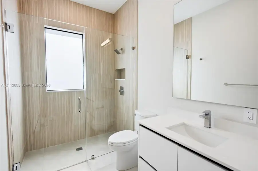 Modern bathroom featuring a glass-enclosed shower with vertical-tile surround, built-in recessed wall niche, wall-to-wall mirror, and white vanity with stone-finish countertop