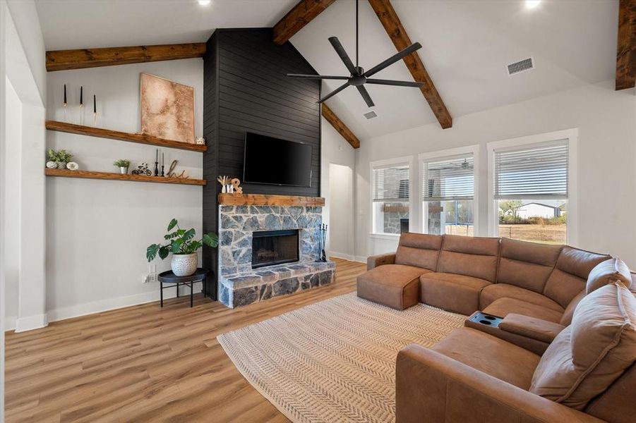 Living room featuring light wood-type flooring, beam ceiling, a stone fireplace, a ceiling fan, and high vaulted ceiling