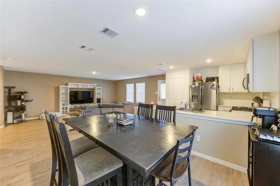 This spacious open-concept living area features a modern kitchen with stainless steel appliances and white cabinetry. The adjoining dining space boasts a large table, perfect for entertaining. The living area includes a comfortable sectional sofa and a built-in entertainment center, creating a cozy atmosphere. Natural light fills the space through multiple windows, enhancing the warm, neutral decor.