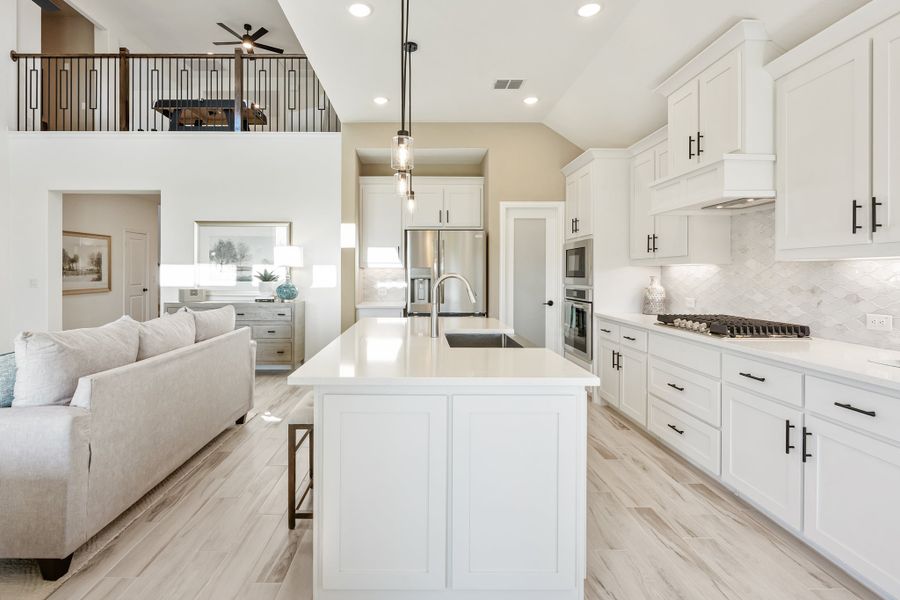 Open-concept kitchen with white cabinets, large island, and stainless appliances, open to living area with second-floor balcony visible above.