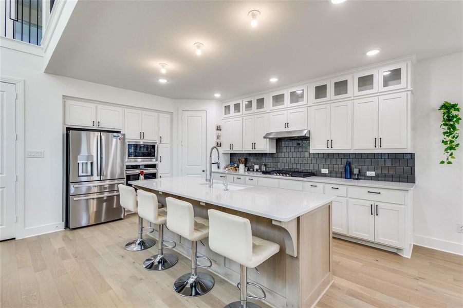 Kitchen featuring stainless steel appliances, a center island with sink, light wood-style floors, white cabinets, and glass fronted cabinets Kitchen featuring stainless steel appliances, a center island with sink, light wood-style floors, white cabinets, and glass fronted cabinets