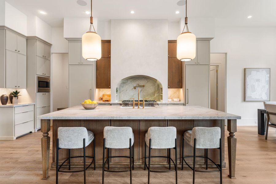 A statement island anchors the kitchen, featuring a substantial countertop with seating and custom wood detailing below. Designer pendant lighting highlights the scale of the space, while the arched cooking alcove creates a striking focal point.