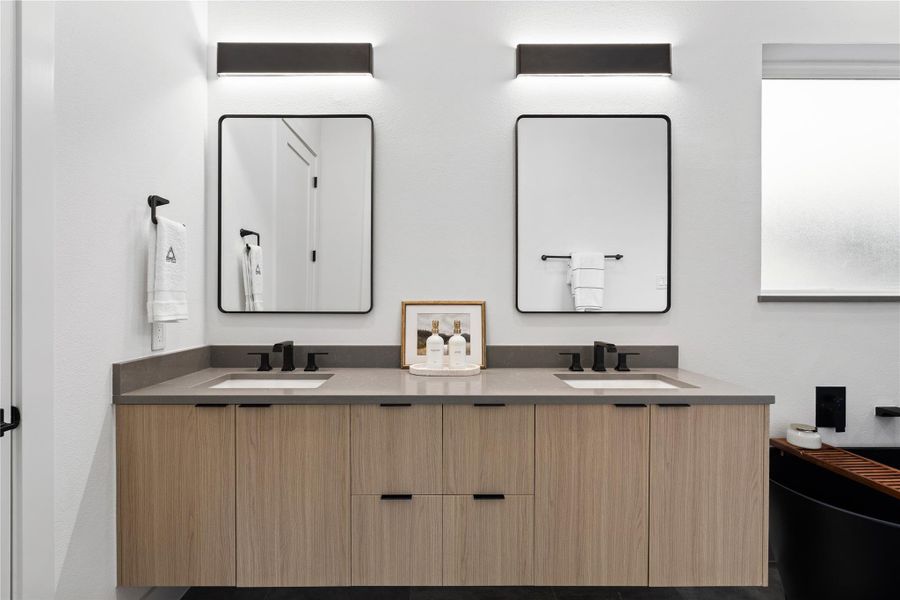 A calm study in symmetry: the primary bath centers a floating, wood-grain vanity with dual under-mount sinks and charcoal quartz surfaces, finished in refined matte-black hardware.Linear sconces and slender framed mirrors provide gallery-level light and proportion, setting a spa-forward tone for unhurried morning rituals.