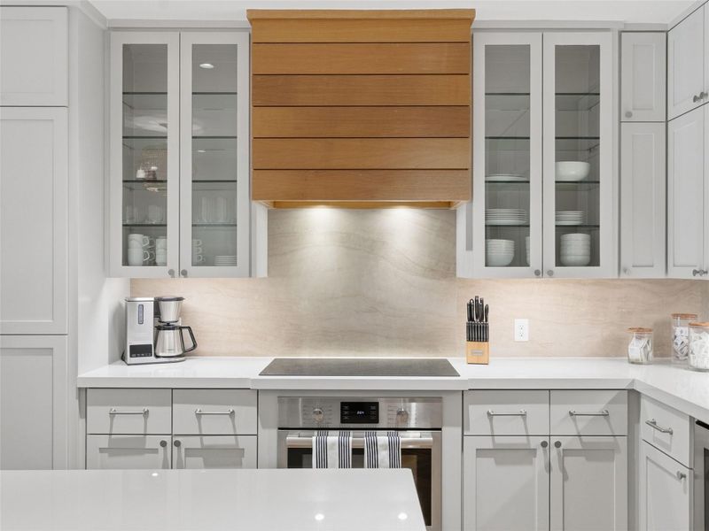Kitchen featuring a wood-panel range hood, glass-front upper cabinets, light-toned backsplash, and a built-in oven Kitchen featuring a wood-panel range hood, glass-front upper cabinets, light-toned backsplash, and a built-in oven