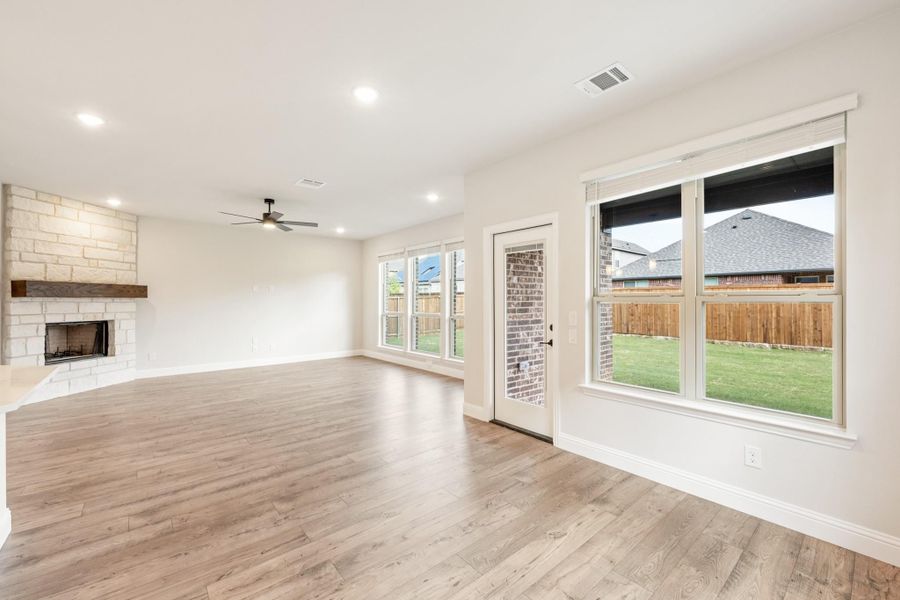 Open living room with stone fireplace, hardwood floors, ceiling fan, and large windows overlooking backyard