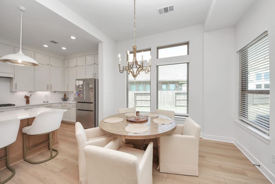 Bright and inviting open-concept kitchen and dining area featuring modern white cabinetry, stainless steel appliances, and a cozy breakfast nook with a stylish chandelier. Large windows provide abundant natural light.