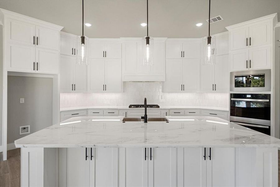 Kitchen featuring quartz countertops, marble tile backsplash, huge island with apron sink & storage cabinets under overhang, ss appliances, undercounter lighting, hidden outlets and pendant lighting.