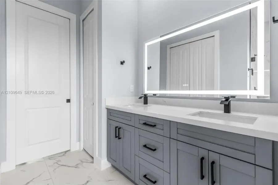 Double vanity with grey shaker-style cabinetry, white solid-surface countertop, integrated dual sinks, matte black fixtures, and a large backlit rectangular mirror
