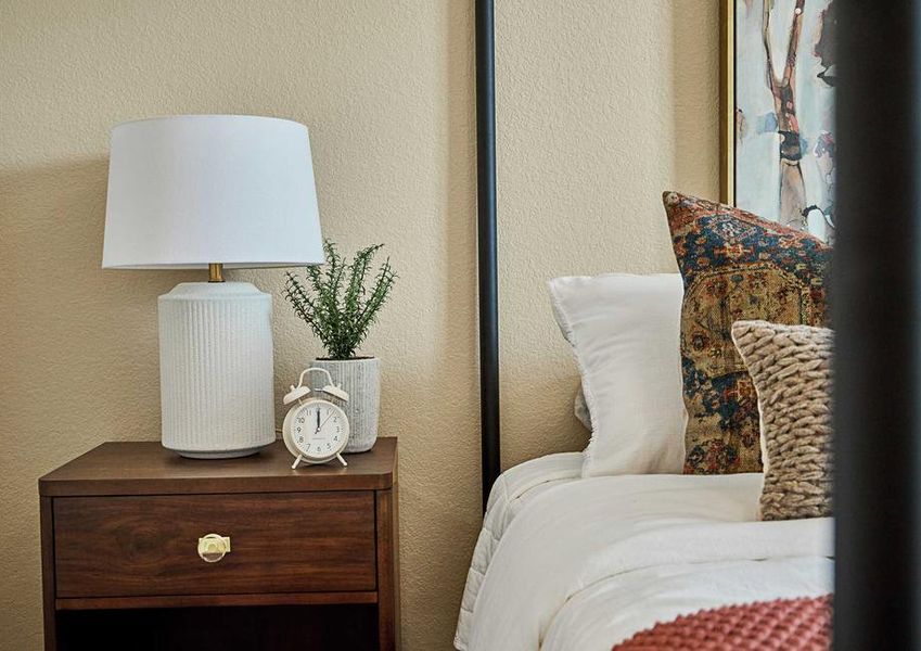 Detail of bed with iron posts, wooden side table with lamp, white sheets and warm natural throw pillows, blanket, nightstand with clock and plant and artwork at head of bed.