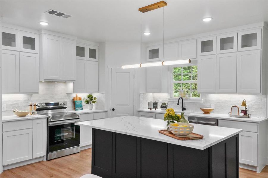 Gourmet kitchen featuring a large central island with a quartz countertop, white shaker cabinetry with some glass-front uppers, stainless steel appliances, and wood-finish flooring Gourmet kitchen featuring a large central island with a quartz countertop, white shaker cabinetry with some glass-front uppers, stainless steel appliances, and wood-finish flooring