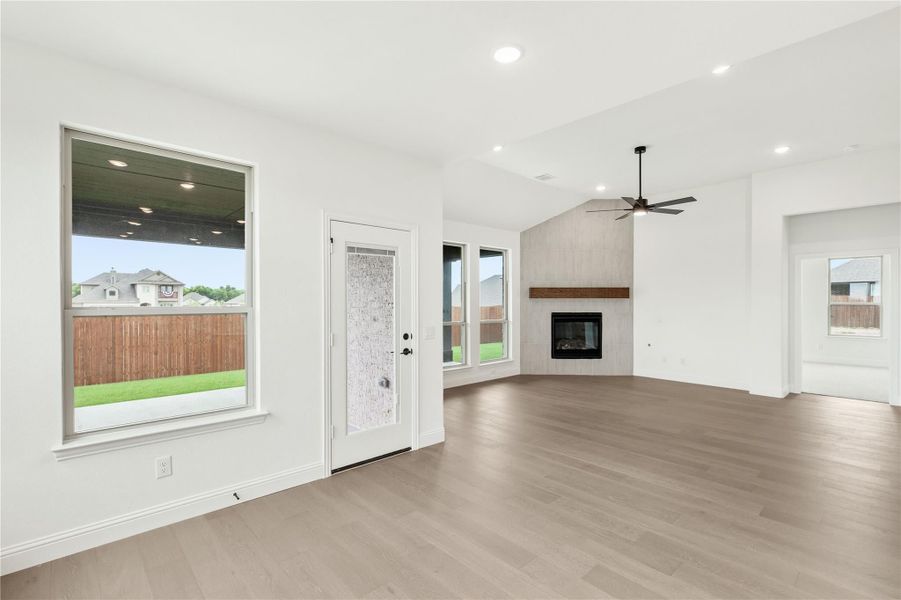 Open living room with light wood floors, ceiling fan, fireplace, and backyard-facing windows Open living room with light wood floors, ceiling fan, fireplace, and backyard-facing windows