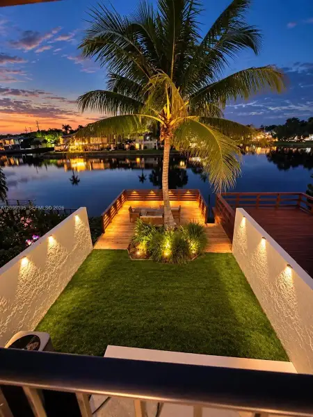 Waterfront property featuring a manicured lawn, wood-plank deck, integrated wall-mounted accent lighting, and a mature palm tree