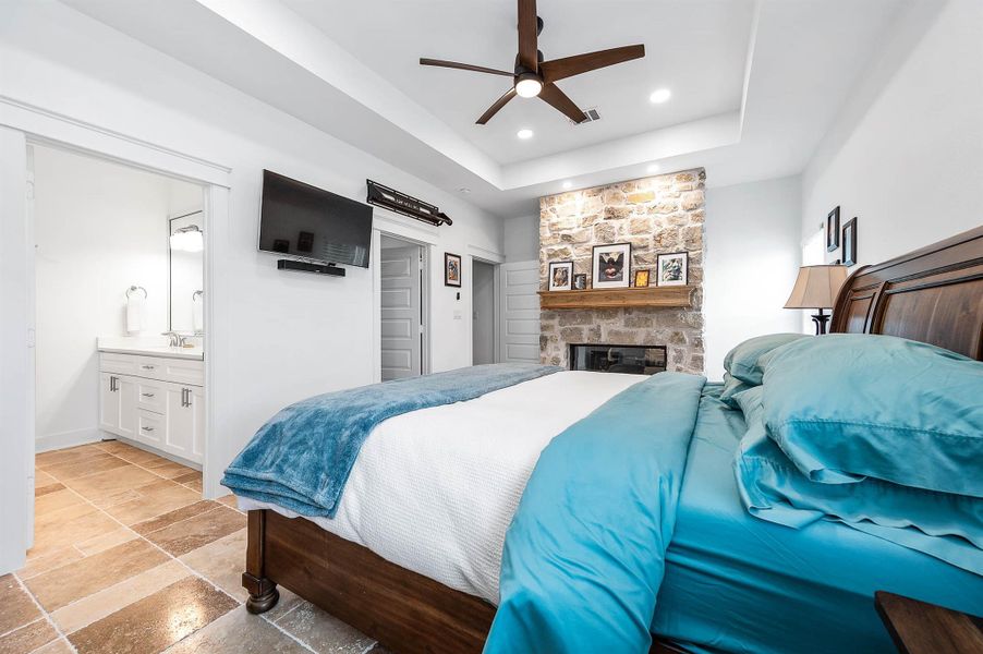 Another view of the spacious primary suite showcasing the cozy stone fireplace, mounted TV, and private en-suite bath access. Recessed lighting, modern finishes, and a ceiling fan add both comfort and style to this relaxing retreat.
