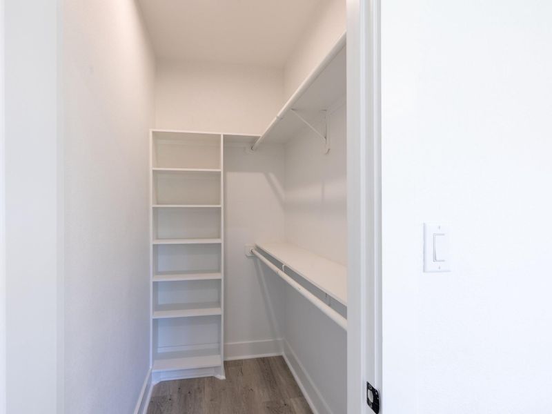 A spacious walk-in closet featuring white walls, built-in shelving, and a long hanging rod for ample storage. The light wood flooring adds a touch of elegance.