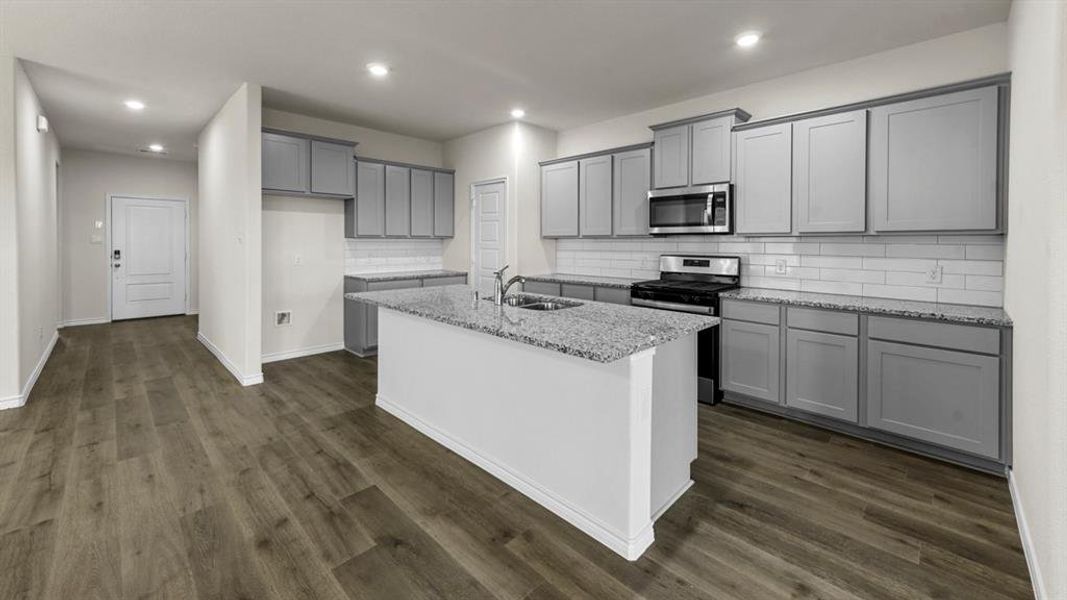 This kitchen features an island with a sink, granite countertops, gray cabinetry, and a subway tile backsplash