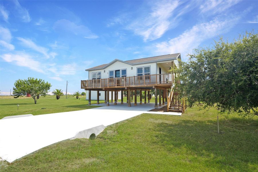 Elevated home with a spacious wooden deck, set on a large, grassy lot. Features a concrete driveway and a stairway leading to the main entrance. Surrounded by open greenery and a few trees, offering a serene atmosphere.