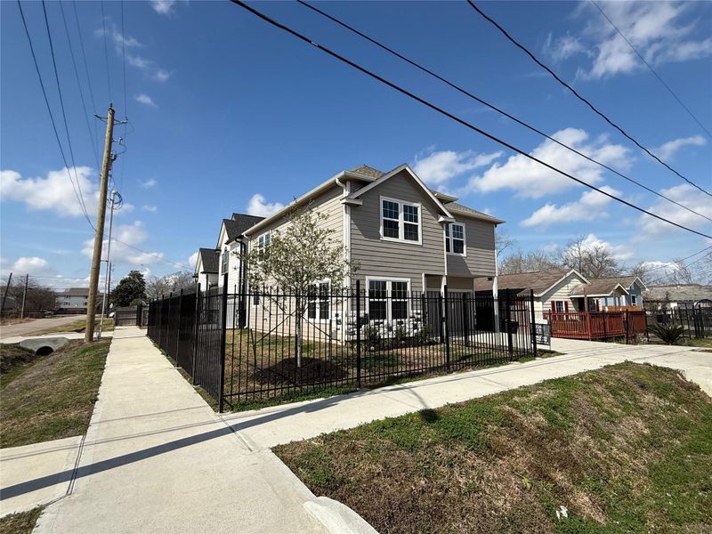 This photo showcases a modern two-story home with light siding, large windows, and a black metal fence. It's situated on a corner lot with a well-maintained sidewalk and some greenery, offering a spacious and inviting exterior.