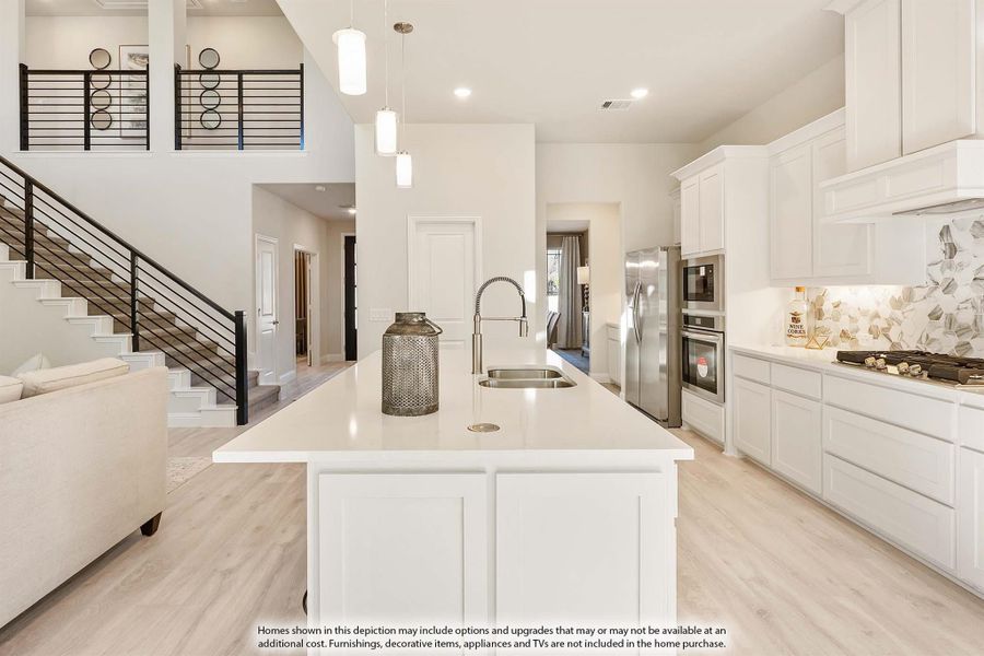 Open kitchen with white cabinets, large island, stainless appliances, and decorative tile backsplash, open to staircase and living area.