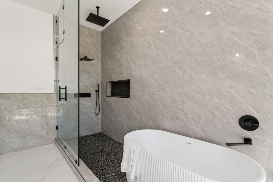 Inside the shower, every detail holds — a ceiling-mount rain head, wall-mounted hand-held fixture, built-in niche at shoulder height, and matte black hardware throughout, all against floor-to-ceiling large-format gray tile. The pebble tile floor and the freestanding tub visible just beyond the glass complete a primary bath that earns every superlative.