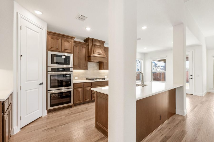 Kitchen with brown wood cabinets, double wall ovens, and a white island with sink on light hardwood floors
