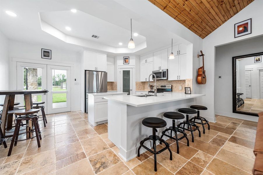 This beautifully designed kitchen features crisp white cabinetry, stainless steel appliances, and a large island with bar seating, creating both style and functionality. Natural light pours in through the expansive windows and glass doors, while the wood-accented ceiling adds warmth and character to the space.