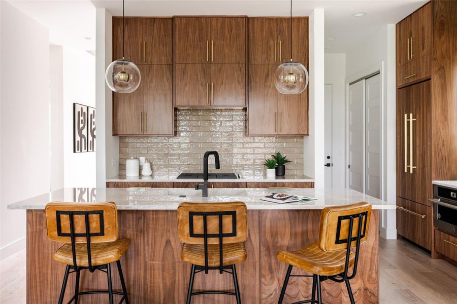 A central island with a sleek sink anchors the kitchen, illuminated by elegant glass globe light fixtures. Warm wood cabinets are accented with champagne-colored pull handles, adding sophistication.