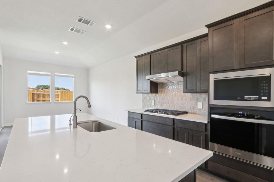 Kitchen featuring tasteful backsplash, appliances with stainless steel finishes, dark brown cabinets, light stone counters, and an island with sink Kitchen featuring tasteful backsplash, appliances with stainless steel finishes, dark brown cabinets, light stone counters, and an island with sink