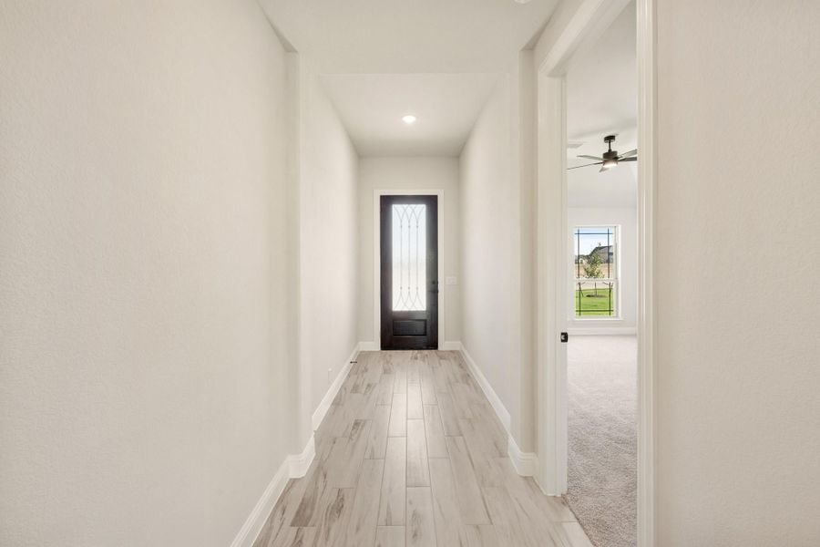 Narrow hallway with light wood floors leading to a dark front door with glass panel insert