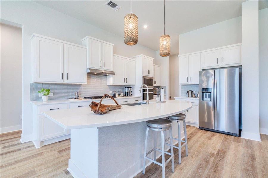 Kitchen featuring stainless steel appliances, white cabinets, backsplash, decorative light fixtures, and an island with sink Kitchen featuring stainless steel appliances, white cabinets, backsplash, decorative light fixtures, and an island with sink