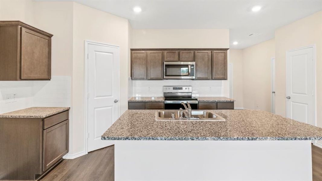 Kitchen featuring an island with an undermount sink, granite-style countertops, wood-finish flooring, dark wood cabinetry, and a subway tile backsplash