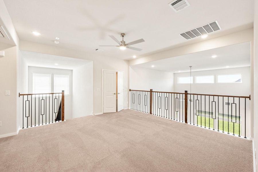 Open upstairs loft with carpet flooring, ceiling fan, and metal railing along open staircase and overlook Open upstairs loft with carpet flooring, ceiling fan, and metal railing along open staircase and overlook