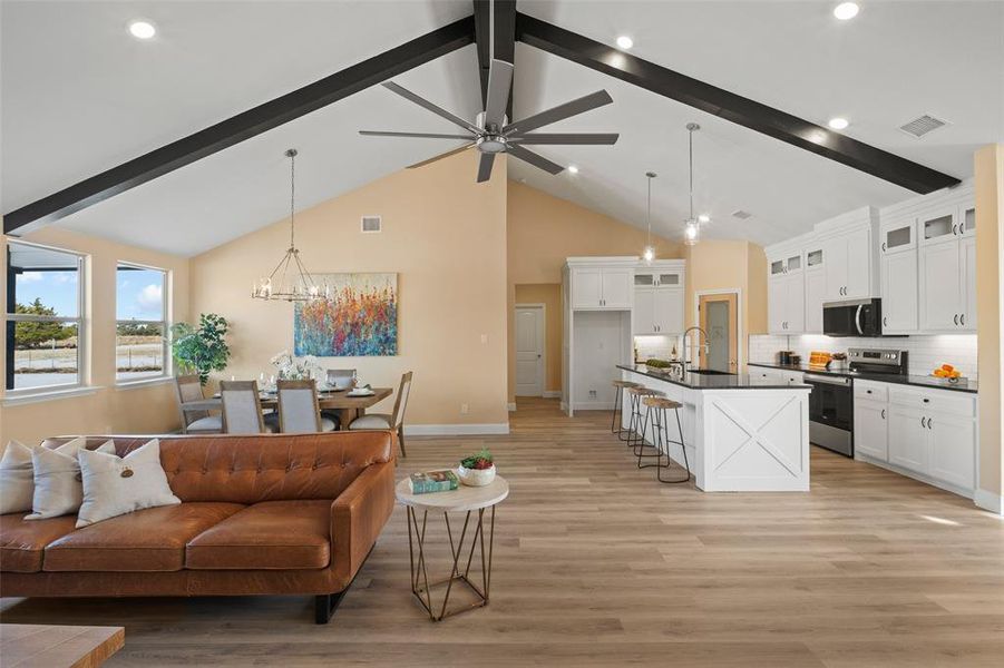 Expansive open-concept living space featuring vaulted ceilings with exposed beams, wood-finish flooring, and white kitchen cabinetry with dark countertops Expansive open-concept living space featuring vaulted ceilings with exposed beams, wood-finish flooring, and white kitchen cabinetry with dark countertops