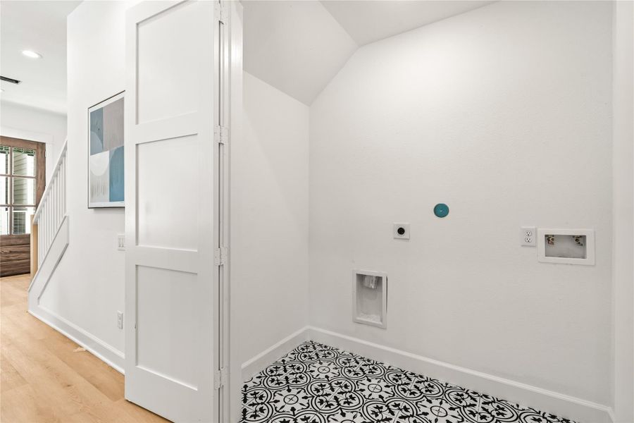 A dedicated laundry closet nestled under the stairs, showcasing graphic black-and-white patterned floor tile, standard utility hookups, and a white bifold privacy door. A dedicated laundry closet nestled under the stairs, showcasing graphic black-and-white patterned floor tile, standard utility hookups, and a white bifold privacy door.