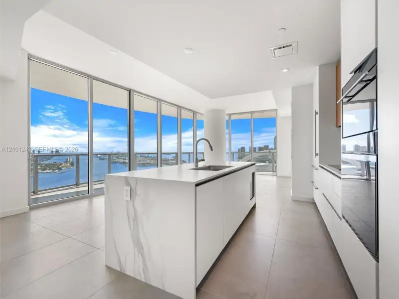 Kitchen featuring a central island with stone-finish waterfall edges, stainless steel under-mount sink, integrated appliances, floor-to-ceiling windows, and tile flooring Kitchen featuring a central island with stone-finish waterfall edges, stainless steel under-mount sink, integrated appliances, floor-to-ceiling windows, and tile flooring