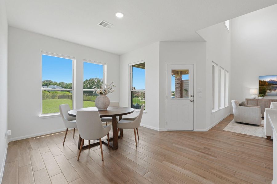 This photo showcases a bright and airy open-concept dining and living area. It features large windows offering plenty of natural light and views of a green backyard. The space has a modern, clean design with wood-like tile flooring and neutral tones, creating a welcoming atmosphere.