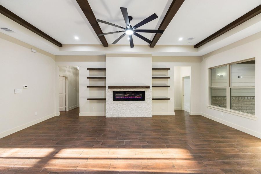 Spacious living room with modern design, featuring a central electric fireplace with stone surround, dark wood ceiling beams, and built-in shelving. Large windows provide natural light, complementing the sleek tile flooring.