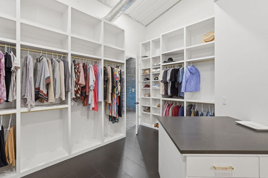 This photo showcases a spacious, organized walk-in closet with ample shelving and hanging space for clothes, hats, and accessories. It features a sleek island with drawers, modern decor, and a well-lit, open design.