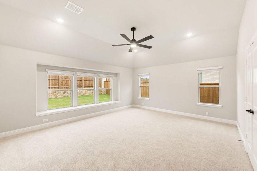 Spacious empty bedroom with beige carpet, ceiling fan, and multiple windows overlooking a fenced backyard Spacious empty bedroom with beige carpet, ceiling fan, and multiple windows overlooking a fenced backyard