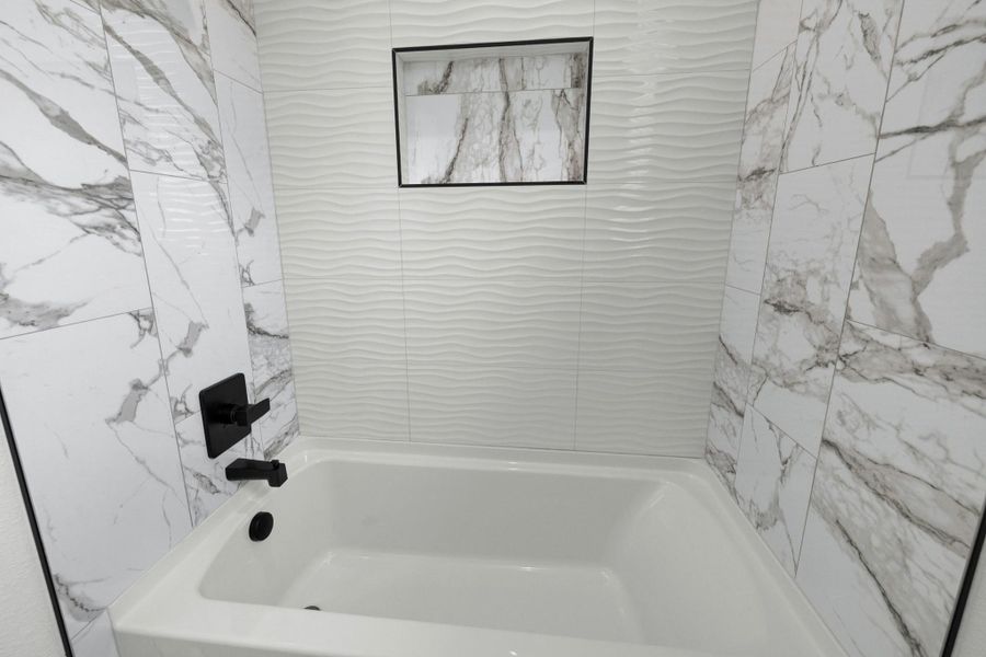 Bathtub with matte black fixtures, surrounded by white marble-look tile and a textured wave-patterned wall