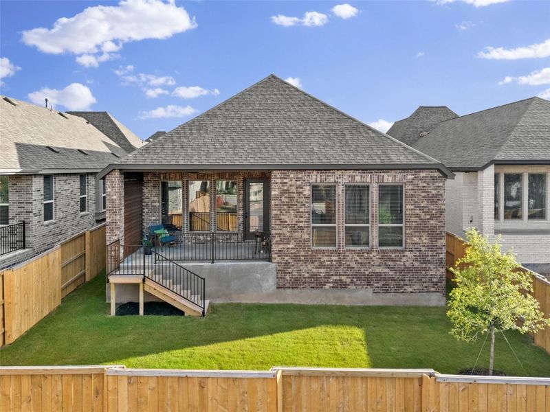 Rear elevation featuring a brick exterior, covered patio with black metal railings, and a fenced backyard with a young tree