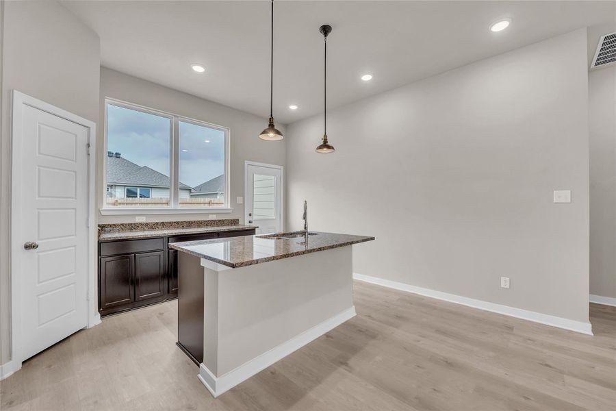 Kitchen with light stone counters, a center island with sink, decorative light fixtures, and light wood-style floors