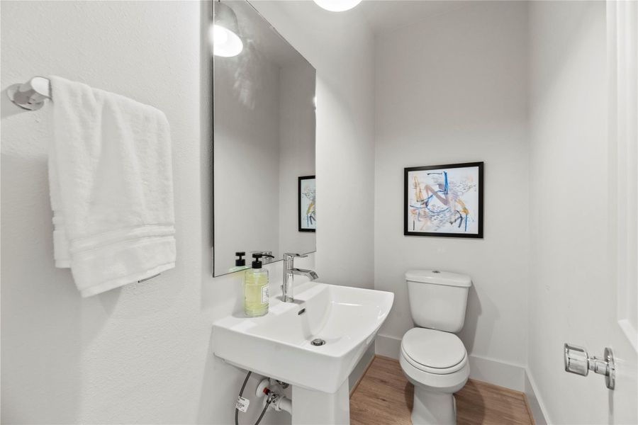 This bright, modern half-bath features a sleek pedestal sink, wall-mounted mirror, and elegant fixtures. Neutral tones, abstract art, and light wood flooring come together to create a clean, inviting space with a stylish touch.