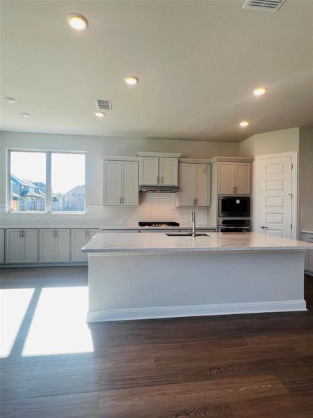 Kitchen with light stone countertops, a center island with sink, backsplash, recessed lighting, and dark wood-style flooring