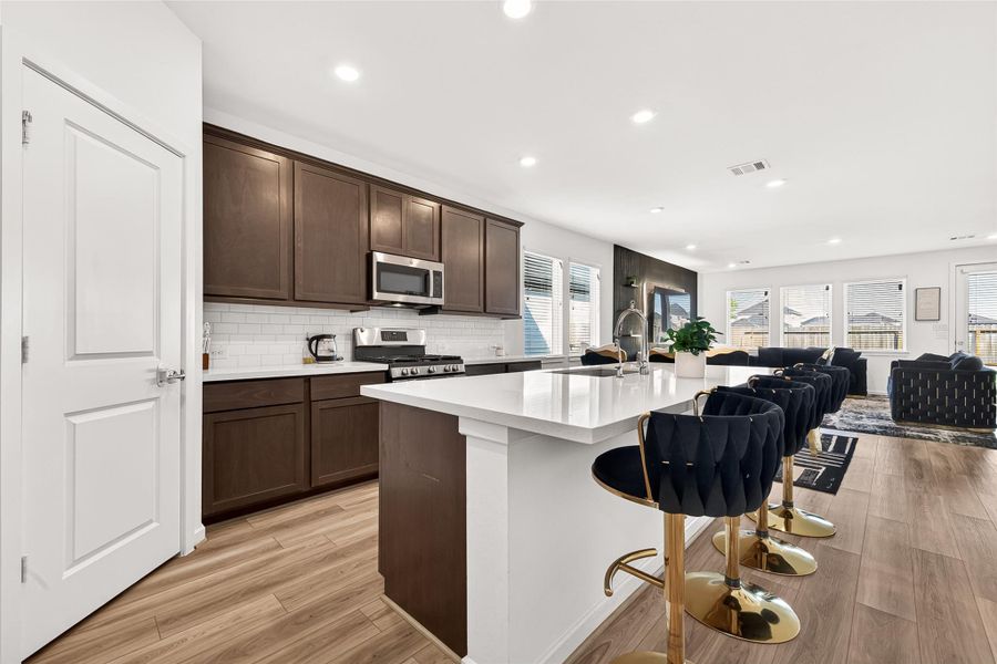 Open-concept kitchen and living area featuring a large quartz island with seating, rich cabinetry, stainless steel appliances, subway tile backsplash, and seamless flow into the main living space. The thoughtfully designed layout is ideal for entertaining while offering a warm, modern aesthetic with abundant natural light throughout.