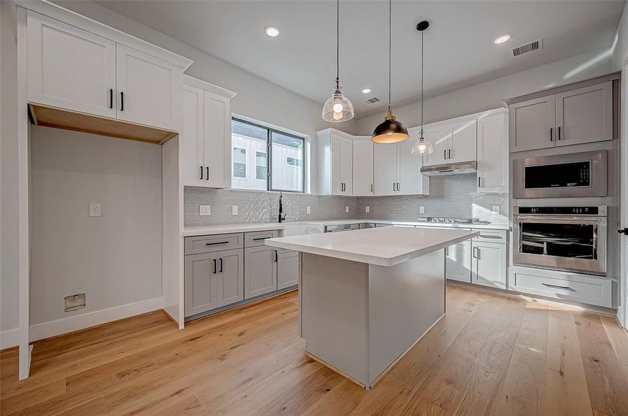 This modern kitchen features sleek cabinetry, stainless steel appliances, and a spacious island with pendant lighting. The light wood flooring and ample natural light create a warm, inviting atmosphere.