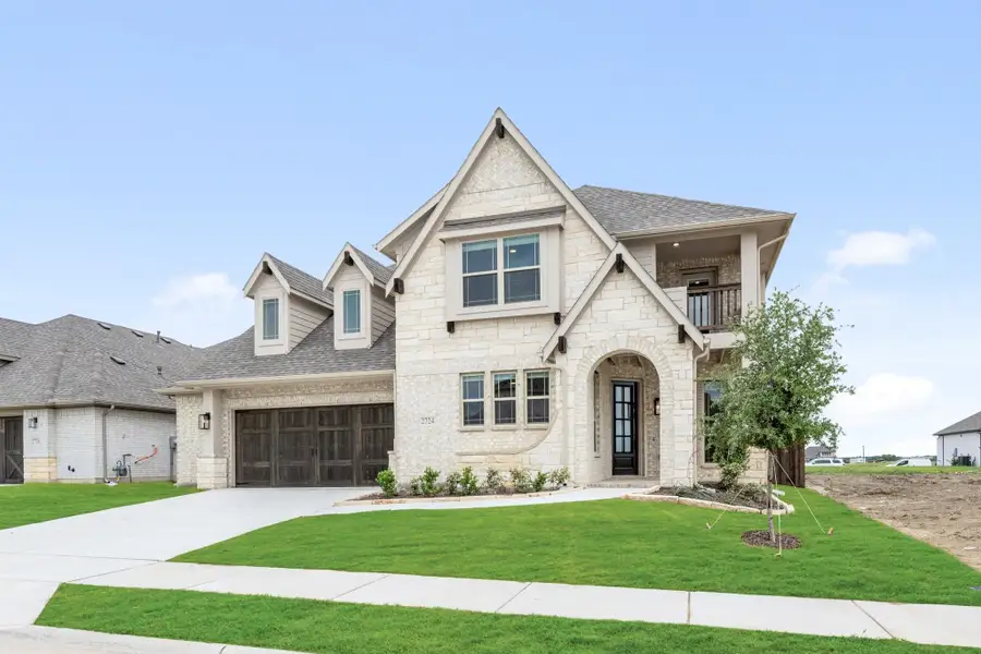 Two-story home with light stone exterior, two-car garage, arched entry, and upper balcony on a corner lot with green lawn