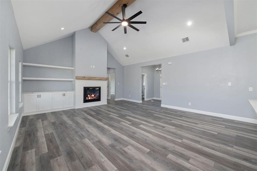 Unfurnished living room with beamed ceiling, dark wood-type flooring, high vaulted ceiling, a glass covered fireplace, and ceiling fan Unfurnished living room with beamed ceiling, dark wood-type flooring, high vaulted ceiling, a glass covered fireplace, and ceiling fan